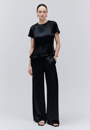 Nő hátul simított hajjal, fekete rövid ujjú szatén felsőt, hozzá illő bőszárú nadrágot, karika fülbevalókat és fekete nyitott orrú szandált visel, áll.