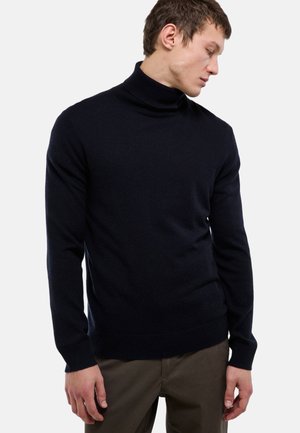 Jeune homme portant un pull à col roulé noir et un pantalon marron, regardant vers le bas et sur le côté sur un fond uni.