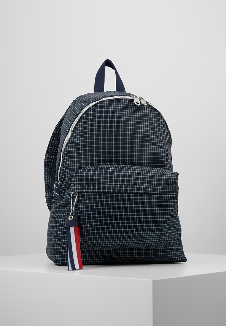 Sac à dos bleu marine avec un motif en quadrillage, fermeture éclair, poche avant et une étiquette rouge, blanche et bleue. Fabriqué en tissu durable avec une finition texturée.