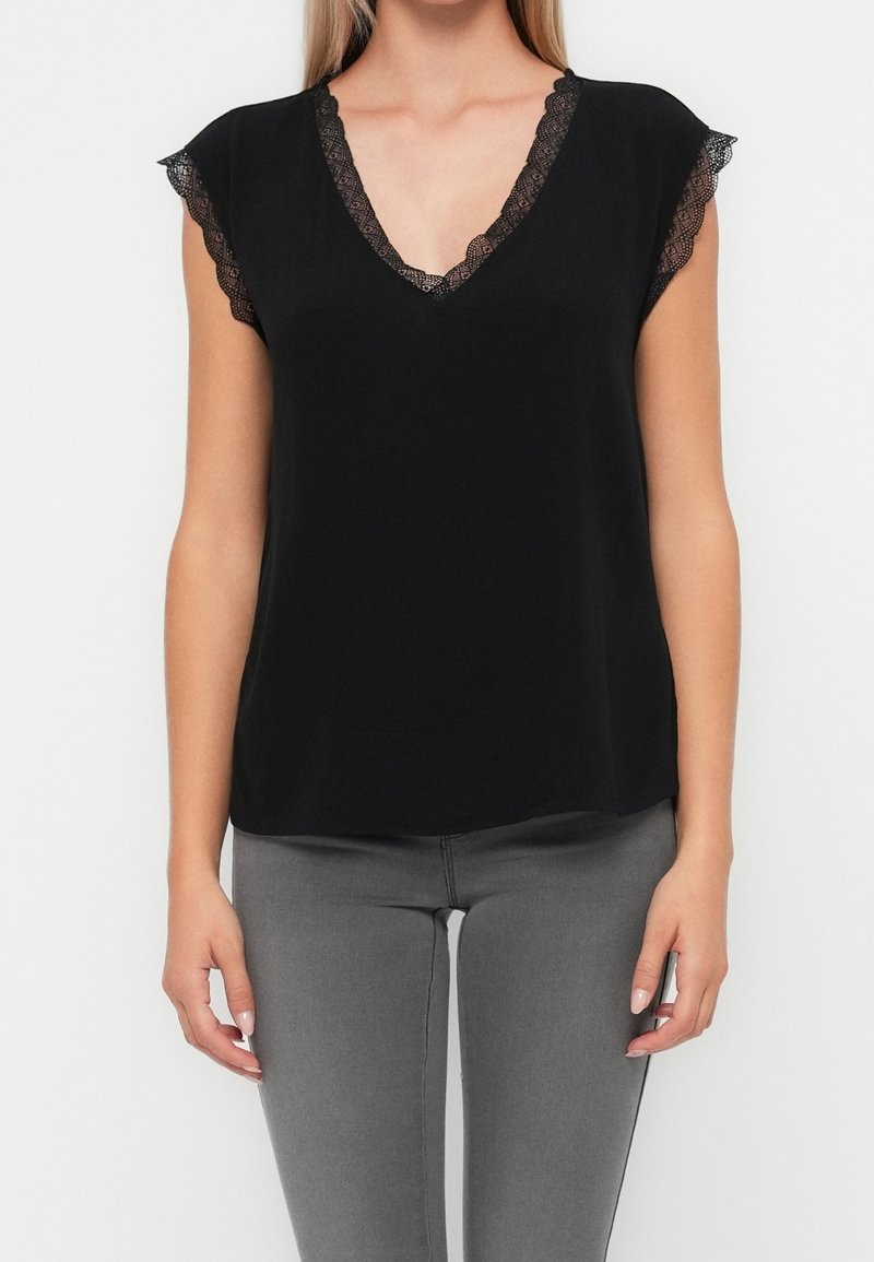 Femme portant un haut noir sans manches à encolure en V avec une bordure en dentelle aux manches et à l'encolure, associé à un pantalon gris.