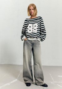 Kobieta z krótkimi blond włosami, ubrana w czarno-białą sweter w paski z napisem "92" oraz szerokie szare dżinsy, stojąca z rękami w kieszeniach, na tle gładkiej ściany.