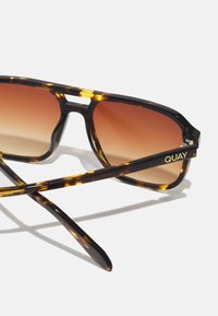 Gafas de sol carey con lentes marrones degradados. Presentan un marco elegante, un puente con un corte sutil y el logo "QUAY" en dorado en la patilla.