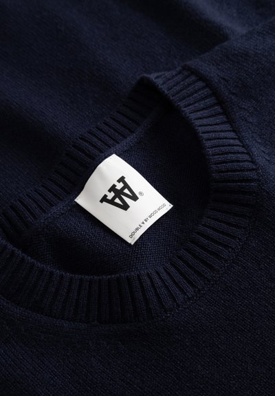 Navy blue knitted sweater featuring a ribbed crew neckline and a fabric label with black text indicating brand and product details.