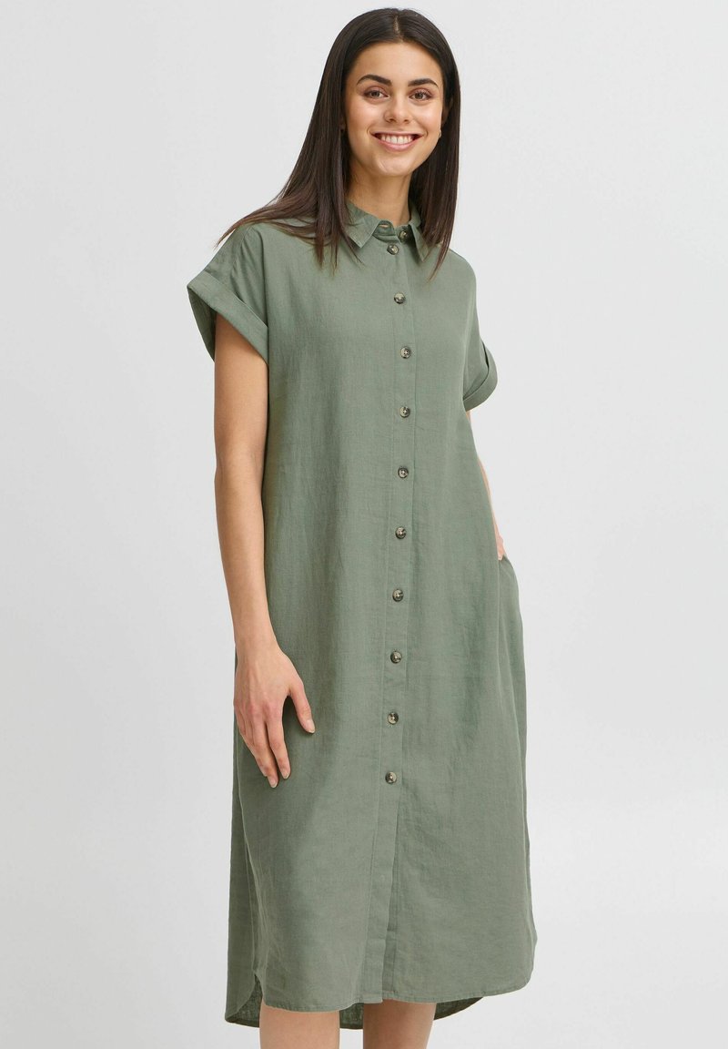 Abito camicia in lino verde khaki con maniche corte arrotolate, bottone frontale e tasche laterali. Presenta una vestibilità rilassata e un orlo fino al ginocchio.