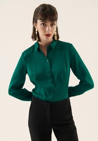 Blusa verde smeraldo a maniche lunghe in tessuto liscio con colletto e chiusura con bottoni, abbinata a pantaloni neri a vita alta.