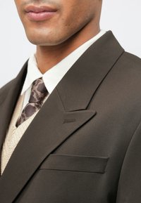 Dark brown tailored blazer with a notch lapel, side pocket, and subtle texture. Worn over a cream collared shirt and patterned tie.