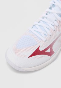 Άσπρο αθλητικό παπούτσι με κόκκινες λεπτομέρειες, με υφή στην επιφάνεια, τμήματα από δίχτυ και επίπεδη κορδόνια. Στρογγυλερή μύτη και ορατή μαλακή σόλα.