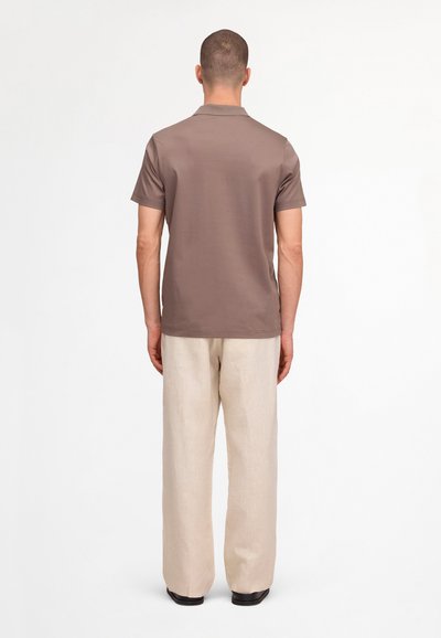 Homme debout de dos, portant une chemise marron à manches courtes, un pantalon beige ample et des chaussures noires sur un fond blanc uni.