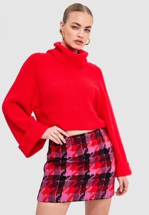 Femme portant un pull à col roulé rouge et une mini-jupe à motifs rouges, roses et noirs, accessoirisée avec des boucles d'oreilles créoles en or et des bagues.