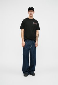 Zwart katoen t-shirt met rode grafische afbeelding, gecombineerd met brede spijkerbroek met blauwe strepen. Gestyled met een zwarte pet en donker schoeisel.