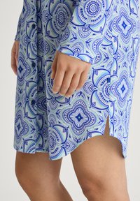 Abito a maniche corte di colore azzurro chiaro con un motivo floreale in blu scuro e verde. Presenta dettagli con bottoni e uno spacco laterale all'orlo.
