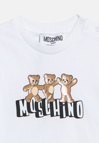 Λευκό βρεφικό μπλουζάκι με τρία χαμογελαστά καφέ αρκουδάκια πάνω από έντονη, στιλιζαρισμένη επιγραφή "MOSCHINO" και ετικέτα στον λαιμό "MOSCHINO BABY".