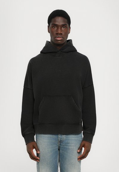 Schwarzer Kapuzenpulli aus weichem Stoff, mit einer Kängurutasche, gerippten Bündchen und einem gestickten Logo auf der Brust. Getragen mit hellblauen Jeans.