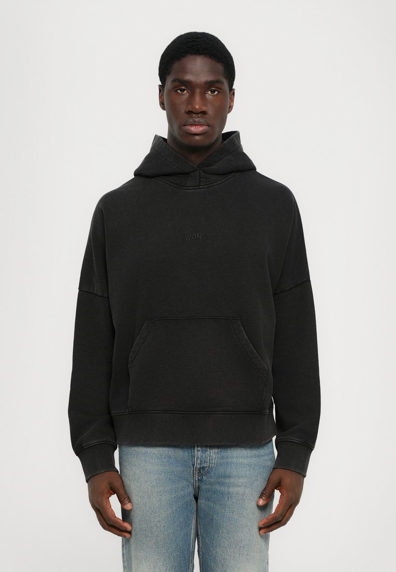 Schwarzer Kapuzenpulli aus weichem Stoff, mit einer Kängurutasche, gerippten Bündchen und einem gestickten Logo auf der Brust. Getragen mit hellblauen Jeans.