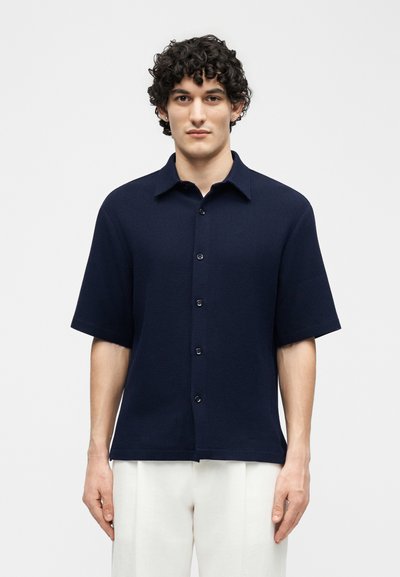 Άνδρας με σγουρά σκούρα μαλλιά, φορώντας μπλε κοντομάνικο πουκάμισο με κουμπιά και λευκό παντελόνι, στέκεται μπροστά σε απλό ανοιχτό φόντο.