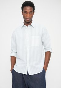 Νεαρός άνδρας με σηκωμένα μανίκια, που φοράει λευκό πουκάμισο με ρίγες και σκούρο μπλε παντελόνι, με τα χέρια στις τσέπες, μπροστά σε απλό φόντο.