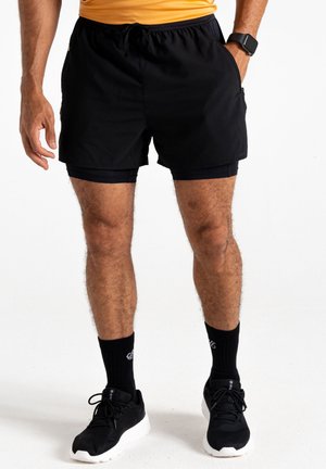 Hombre con pantalones cortos de running negros en capas, calcetines negros, zapatillas de running negras y camiseta amarilla, de pie con una mano en el bolsillo.