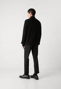 Maglione nero a coste con collo alto, abbinato a pantaloni neri e scarpe. Tessuto liscio e vestibilità rilassata, che enfatizza un look monocromatico.