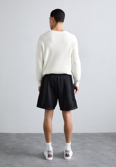 Pull en maille côtelée blanche, shorts noirs et baskets blanches avec des accents colorés en zigzag. Design simple, coupe décontractée et tissu texturé.