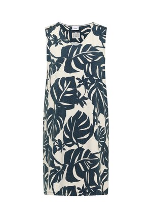 Jeune femme souriante, portant une robe blanche sans manches avec de grands motifs de feuilles tropicales bleu foncé et des chaussures beiges, debout devant un fond uni.