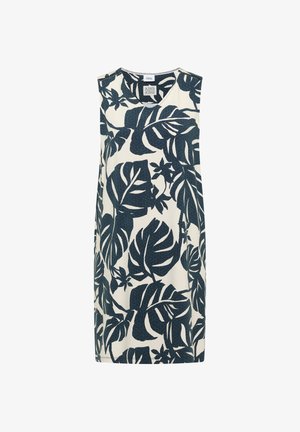 Jeune femme souriante, portant une robe blanche sans manches avec de grands motifs de feuilles tropicales bleu foncé et des chaussures beiges, debout devant un fond uni.