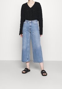 Persona che indossa un maglione nero a maniche lunghe con scollo a V, jeans a gamba larga di colore blu chiaro e sandali neri senza tacco, in piedi contro uno sfondo bianco.