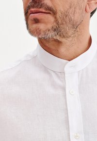Biała lniana koszula z kołnierzykiem mandaryńskim, z subtelną teksturą i listwą z białymi guzikami w okolicy szyi.