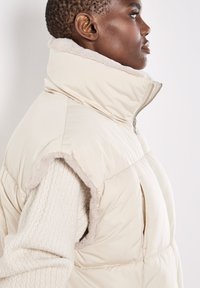 Beige gewatteerde vest met een hoge kraag en zachte faux fur voering. Heeft een gladde buitenkant, ritsluiting en armsgaten. Textuur van de onderlaag zichtbaar.