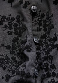 Melnas auduma kardigāns ar teksturētu ziedu izšuvumu, ar melniem pogām un gludu fonu. Tuvplāns uz dizaina un materiāla detaļām.