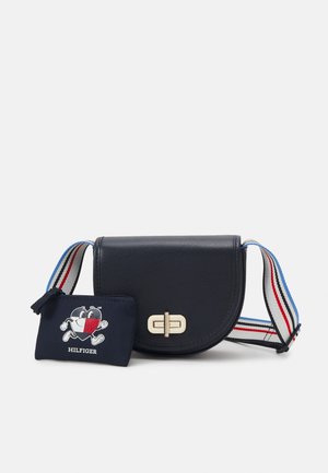 Granatowa torba na ramię z złotą klamrą i paskiem w paski, zestawiona z małą granatową saszetką z kreskówkową twarzą i tekstem "Hilfiger".