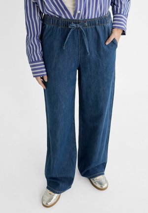 Person wearing loose dark blue denim pants with drawstring waist, a purple and white striped shirt, and silver sneakers, standing with hands partly in pockets.