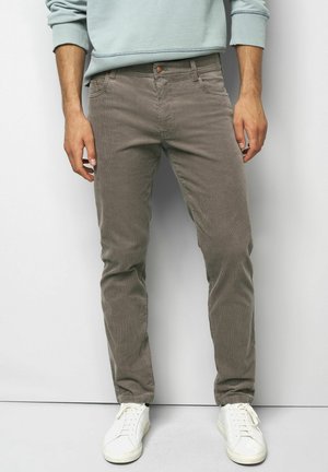 Man wearing taupe corduroy pants, white sneakers, and a light blue sweatshirt standing against a plain white background.