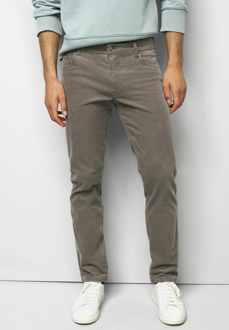 Man wearing taupe corduroy pants, white sneakers, and a light blue sweatshirt standing against a plain white background.
