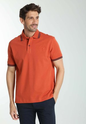 Homme portant un polo orange avec des bordures noires sur le col et les manches, debout avec une main dans la poche, souriant et regardant vers la droite.