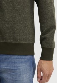 Donkergroen trui met een gestructureerd breipatroon, geribde boorden en onderkant. Gdraag over lichtblauwe denim, wat zorgt voor contrasterende kleuren en materialen.