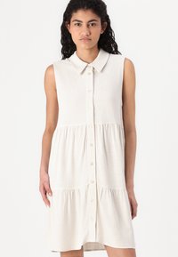 Robe sans manches beige clair avec un col, jupe à volants et texture subtile. Présente des poches avant et une coupe décontractée.