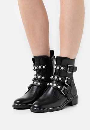 Botas de tobillo de cuero negro con tres correas de hebilla adornadas con perlas y tachuelas. Presentan una puntera redondeada y un tacón bajo apilado.