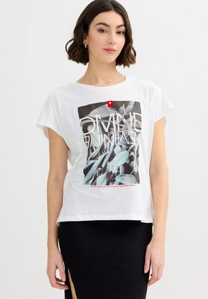 Mujer con cabello corto y castaño, vistiendo una camiseta blanca con gráfico y una falda negra con abertura, de pie frente a un fondo blanco.