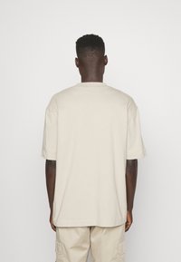 Beige cotton t-shirt with a relaxed fit, short sleeves, and a crew neckline, viewed from the back against a plain white background.