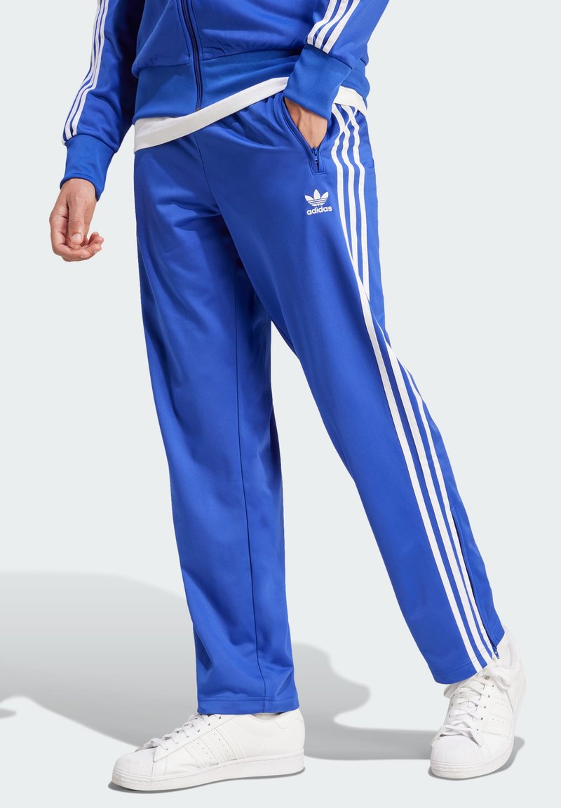 Zilas sporta bikses ar baltām sānu svītrām, izgatavotas no gluda poliestera auduma, ar elastīgu jostasvīru un adidas logotipu.