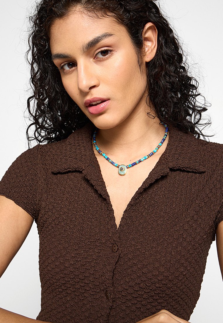 Braunes, strukturiertes Strickhemd mit kurzen Ärmeln und Kragen. Bunte Perlenkette mit einem zentralen Anhänger.