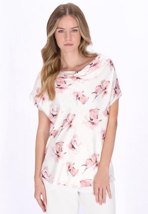 Jeune femme aux longs cheveux ondulés portant un chemisier blanc à motifs floraux roses et un pantalon blanc, debout devant un fond uni.