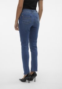 Jeans de mezclilla azul de tiro alto con un corte ajustado, bolsillos traseros con detalles de costura, combinados con tacones altos negros.