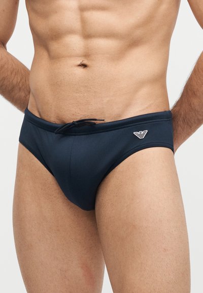 Ανδρικός κορμός που φοράει σκούρα μπλε μαγιό με κορδόνι και μικρό λογότυπο στην αριστερή πλευρά, τα χέρια ακουμπούν πίσω από την πλάτη.