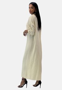 Lang, tekstureret cremefarvet kjole med løs pasform og op rullede ærmer, med brystlomme. Modellen står foran en neutral baggrund.