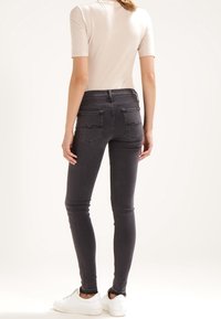 Mujer vestida con jeans ajustados negros, top de manga corta blanco y zapatillas blancas, de pie y mirando hacia el lado en un fondo liso.