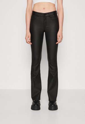 Personne portant un pantalon moulant en similicuir noir et des chaussures à plateforme noires, debout devant un fond blanc uni.