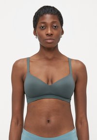 Žena s kratkom kosom nosi prigušeno zelenu bralette i svijetloplave Under Armour kratke hlače, stojeći protiv obične bijele pozadine.