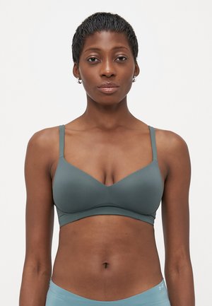 Woman with short hair wearing a muted green bra and light blue Under Armour shorts, standing against a plain white background.