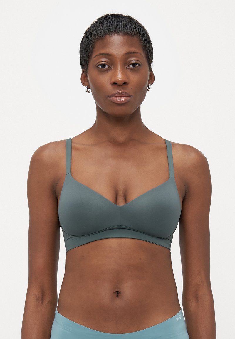 Žena s kratkom kosom nosi prigušeno zelenu bralette i svijetloplave Under Armour kratke hlače, stojeći protiv obične bijele pozadine.